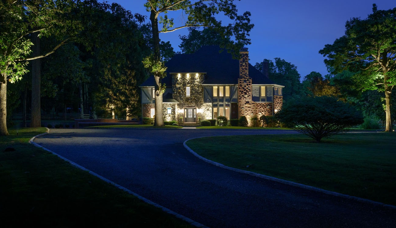 Front Yard Landscape Lighting Tips to Boost Curb Appeal in Mill Neck, NY 3 Front yard landscape lighting highlighting a large stone home, trees, and driveway at dusk.